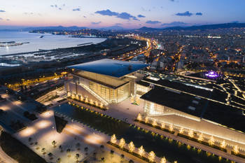 Photo taken with a drone showing the illuminated city at night and the sea focusing on a large glass building surrounded by trees and with a lake in front of it